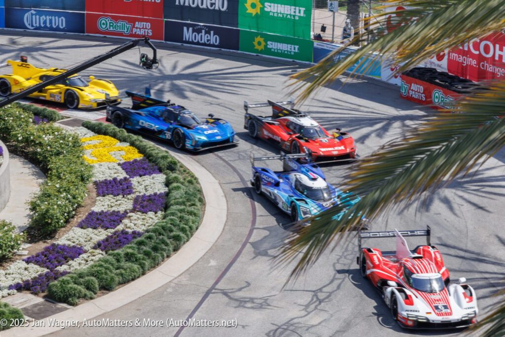 Six sports cars race around a curved track lined with flowers and advertising banners during a daytime motorsport event.