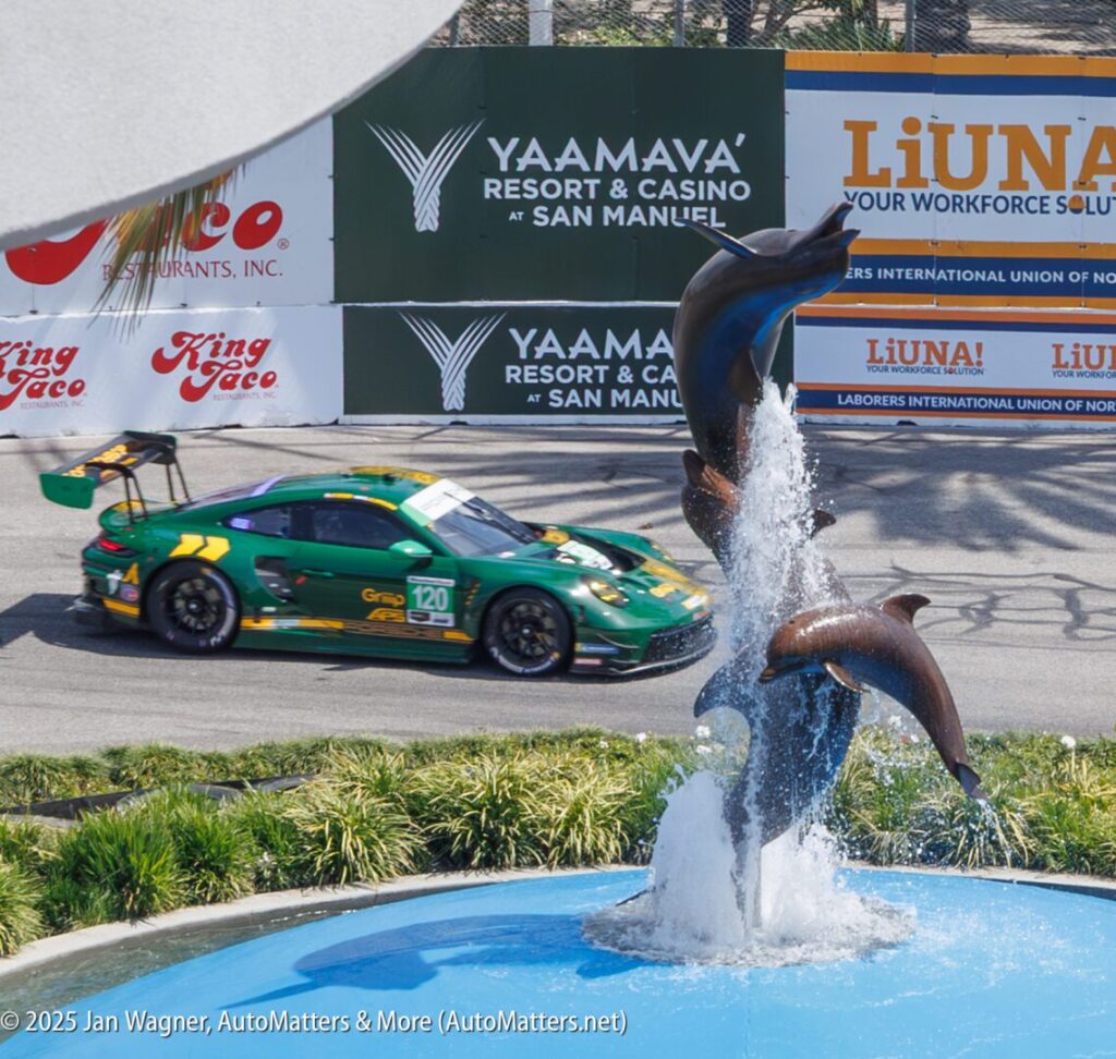 A green race car speeds past a dolphin fountain during a motorsports event, with sponsor banners and palm shadows in the background.