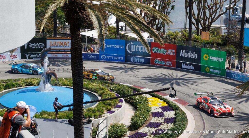 Three race cars navigate a sharp turn on a city street circuit bordered by colorful sponsor banners, with a fountain, palm trees, and flowers in the foreground.