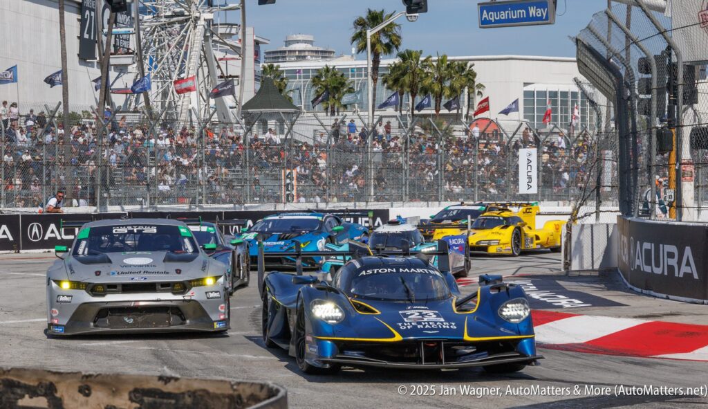 Sports cars race through a turn on a city street circuit during a daytime motorsports event, with spectators and palm trees visible in the background.