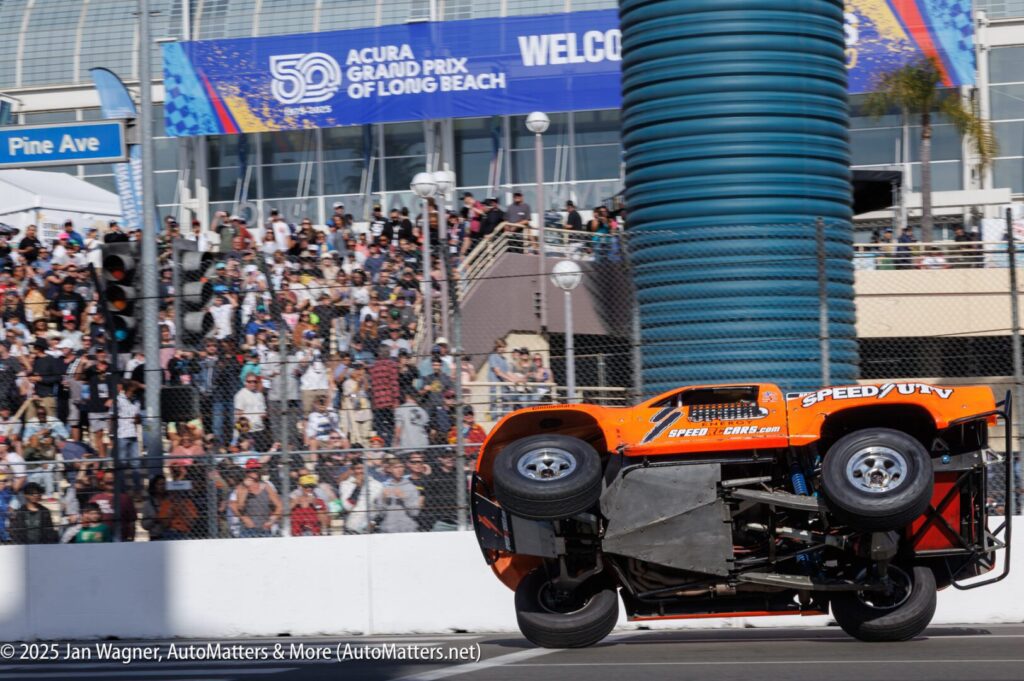 An orange off-road race truck drives on two wheels during a race at the Acura Grand Prix of Long Beach, with a crowd watching from the stands.