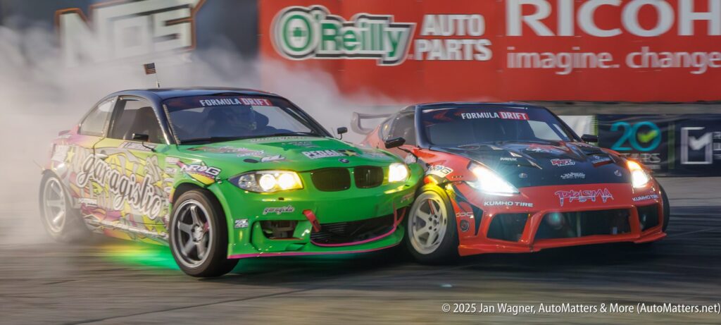 Two modified cars drift side by side on a racetrack, producing tire smoke, with sponsor logos visible on both vehicles.