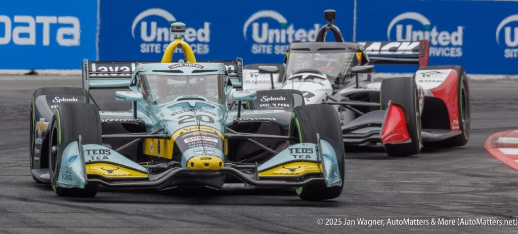 Two IndyCar race cars speed around a track corner; car number 20 leads closely ahead of a black and white competitor.