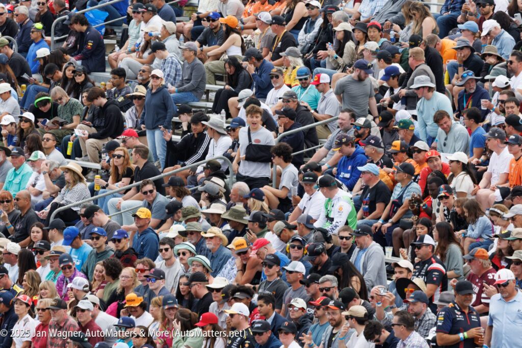 A large crowd of people seated outdoors in stadium-style bleachers, watching an event. Most are wearing hats and sunglasses.