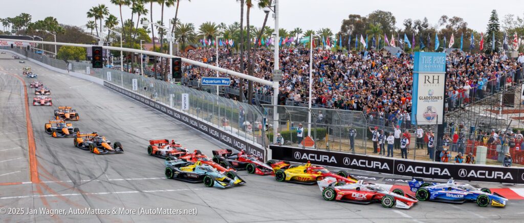 IndyCar race cars take a tight turn on a street circuit as a large crowd watches from the stands and sidewalks lined with palm trees.