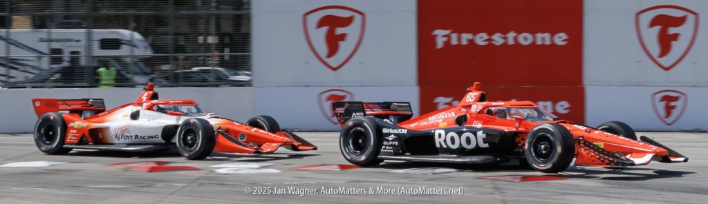 Two red IndyCar race cars drive side by side on a racetrack, passing Firestone-branded barriers during a motorsport event.