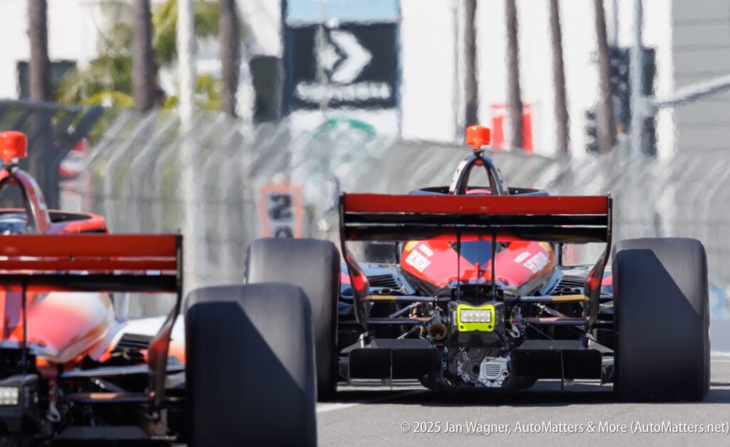 Two open-wheel race cars drive on a track, viewed from behind, with fencing and blurred signage in the background.