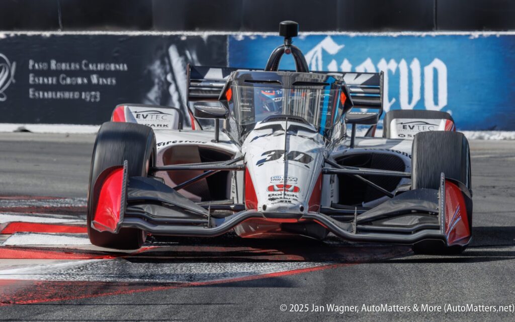 A black and white IndyCar race car, number 30, drives over a red and white curb on a racetrack, with advertisements visible in the background.
