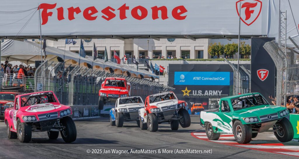 Several off-road race trucks jump and turn on a city street circuit during a stadium super trucks race under a Firestone banner.