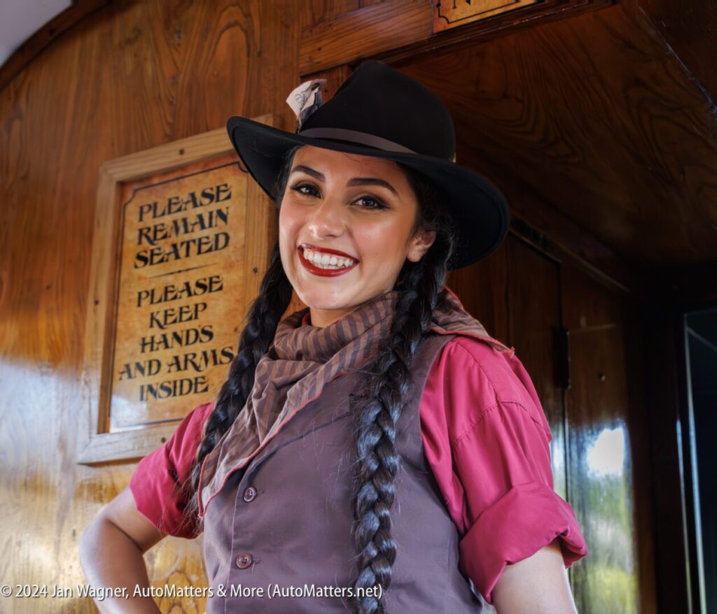 Woman in western-style costume with braided hair and hat smiles inside a wooden train car, standing near signs that read "Please remain seated" and "Please keep hands and arms inside.