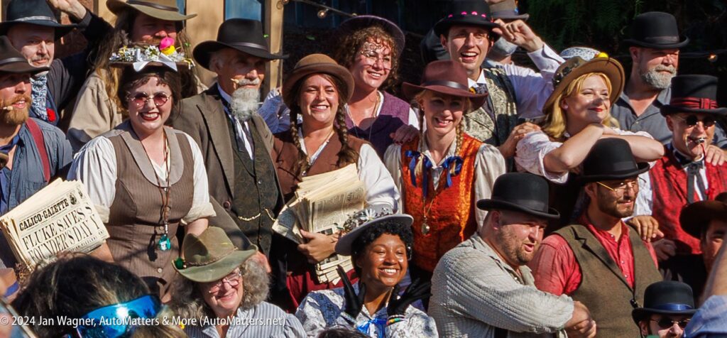 A group of people dressed in Old West costumes pose outdoors, some smiling and holding newspapers, with hats and vests as part of their attire.