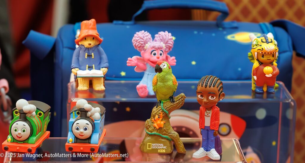 Six colorful children's toy figurines, including characters from Thomas & Friends, Paddington Bear, Trolls, and a National Geographic Kids parrot, displayed in front of a blue bag.