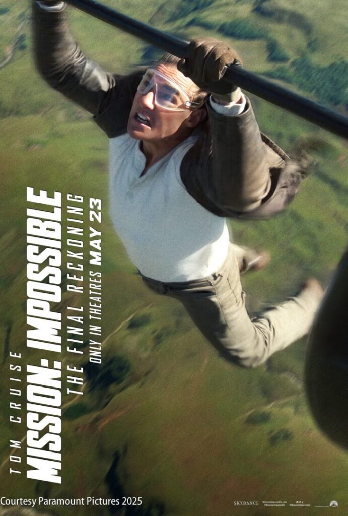 A man wearing glasses and a harness hangs from a moving helicopter over a landscape, with text promoting "Mission: Impossible – The Final Reckoning," releasing May 23, 2025.