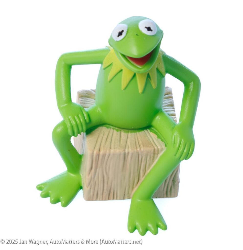 A green frog character figurine with a wide mouth sits on a textured, tan block with its arms resting on its knees.