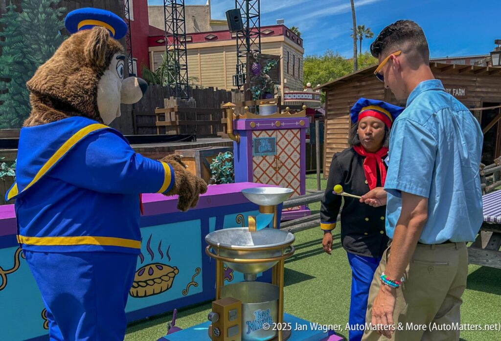 A person in blue hits a ball with a mallet at an outdoor game booth, supervised by a performer and a bear mascot in sailor costumes.