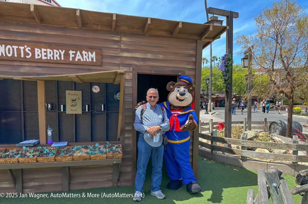 A man stands smiling next to a costumed bear character in front of a wooden Knott’s Berry Farm booth on a sunny day.