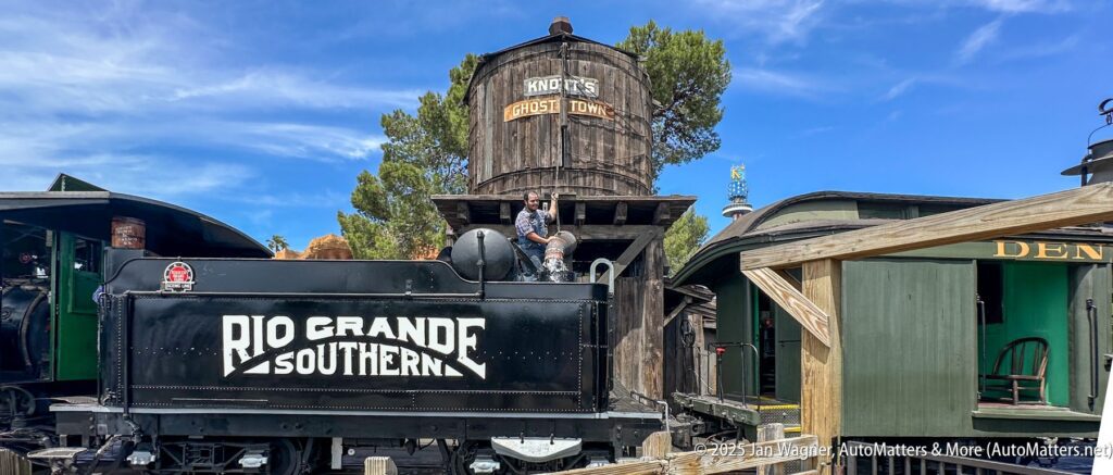 A man sits on a black Rio Grande Southern train engine in front of a wooden water tower labeled "Ghost Town" on a sunny day.