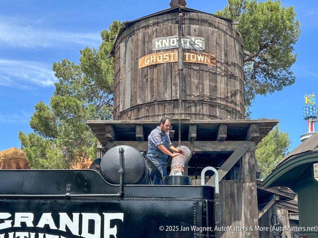 A man stands on a black train engine, filling its water tank from a large wooden water tower labeled "Knott's Ghost Town," with trees and blue sky in the background.