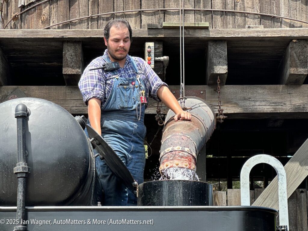 A man in overalls fills a black steam locomotive with water using a large spout at a wooden water tower.