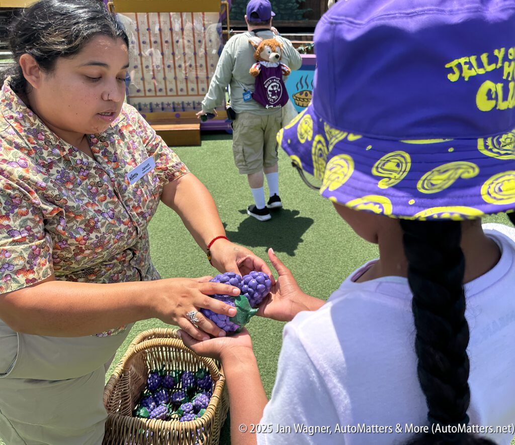 A woman hands a plastic bunch of grapes to a child in a purple hat at an outdoor event, with a basket of similar grapes nearby.