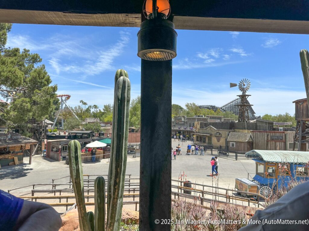 A view of an Old West-style theme park area with cacti in the foreground, visitors walking, rustic buildings, and a windmill in the background under a clear sky.