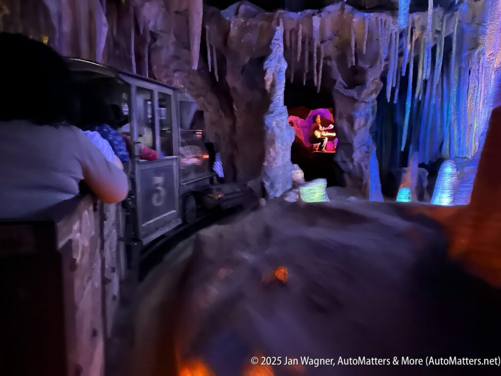 Passengers ride a dark, cave-themed amusement park attraction train, passing colorful illuminated rock formations and a lit animated figure in the distance.