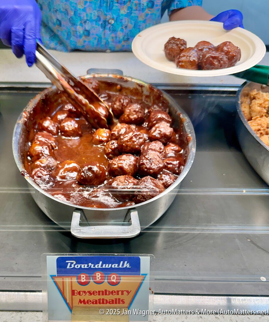 A person wearing gloves serves saucy boysenberry meatballs from a large pan into a plate at a food counter labeled "Boardwalk BBQ Boysenberry Meatballs.
