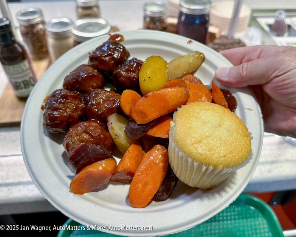 A hand holds a plate with glazed meatballs, roasted carrots and potatoes, and a plain muffin. Condiments and jars are visible in the background.