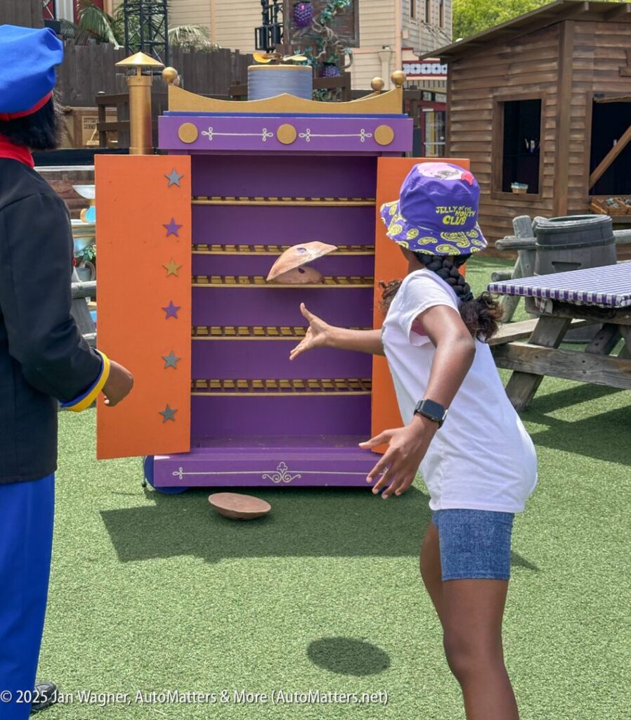 A girl in a purple hat throws a disc toward a colorful board with horizontal slots at an outdoor activity area.