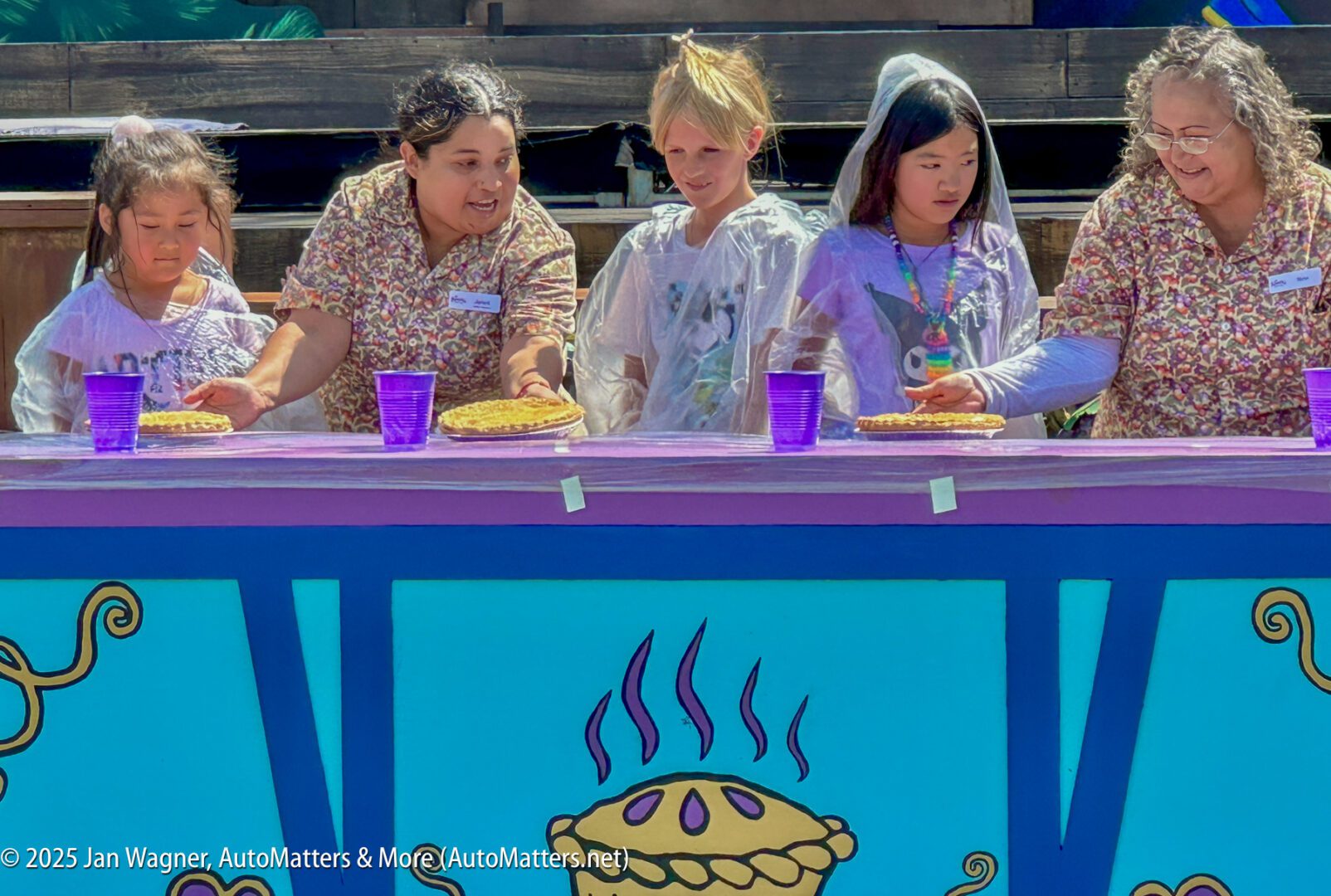 Four people, two adults and two children, stand behind a table with pies and purple cups, all wearing patterned shirts and plastic ponchos, preparing for a pie-eating event.