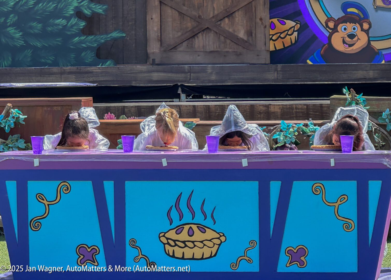 Three people in ponchos compete in a pie-eating contest at an outdoor event, heads down over pies on a decorated blue table.