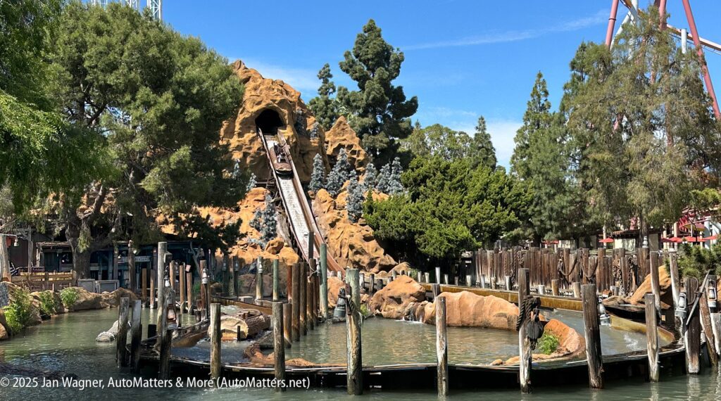 A water log ride descends from an artificial mountain surrounded by trees, with a splash pool and wooden fencing in the foreground.
