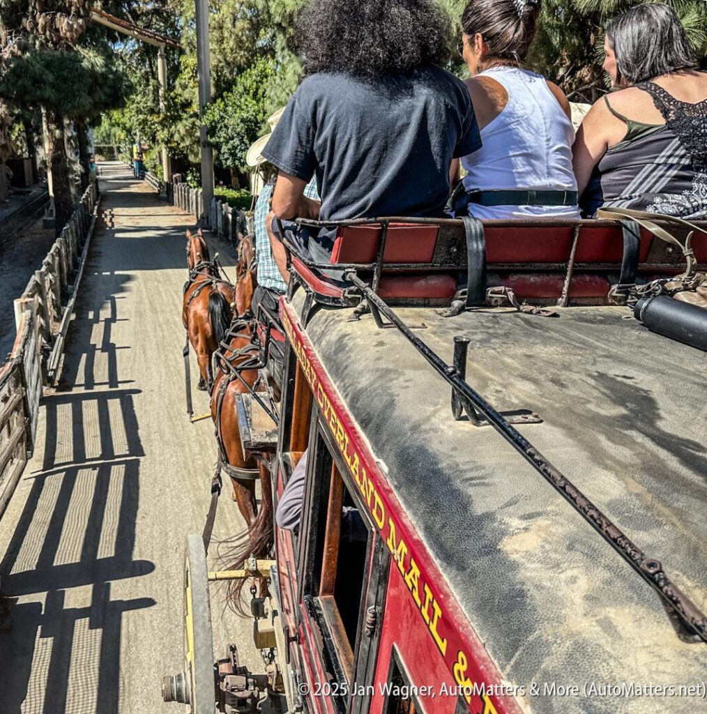 A group of people ride atop a horse-drawn stagecoach on a dirt road beside a wooden fence and trees.