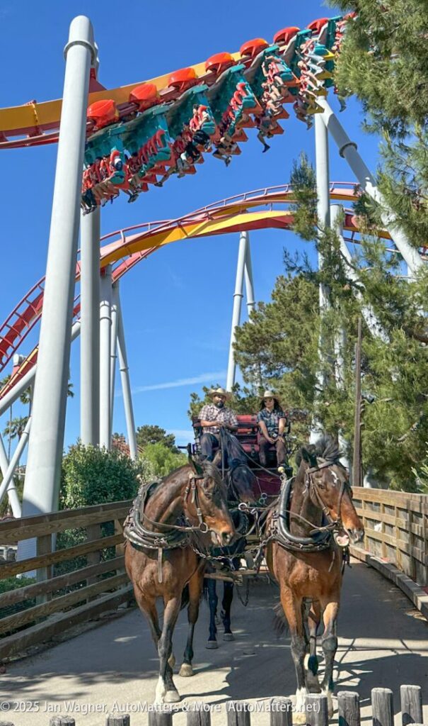 Two horses pull a carriage with two people on a wooden path as a roller coaster speeds overhead against a clear blue sky.