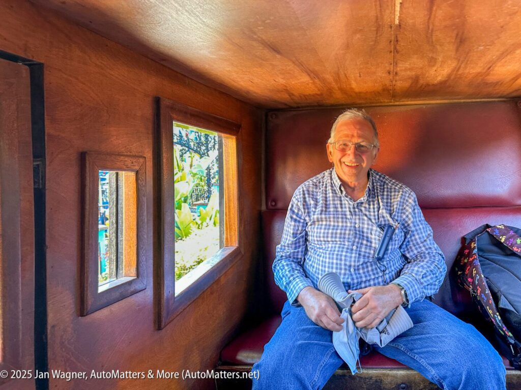 An older man sits and smiles inside a wooden train car, holding a rolled-up newspaper on his lap. Sunlight enters through the windows beside him.
