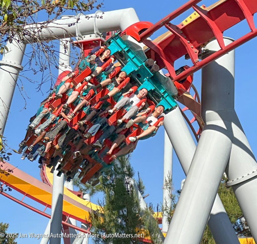 People ride an inverted roller coaster with red tracks and support beams, passing through a curve on a clear day with trees in the background.