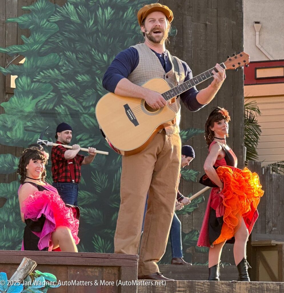 A man in a vest and cap plays an acoustic guitar on an outdoor stage, with performers in colorful costumes and two men holding axes in the background.