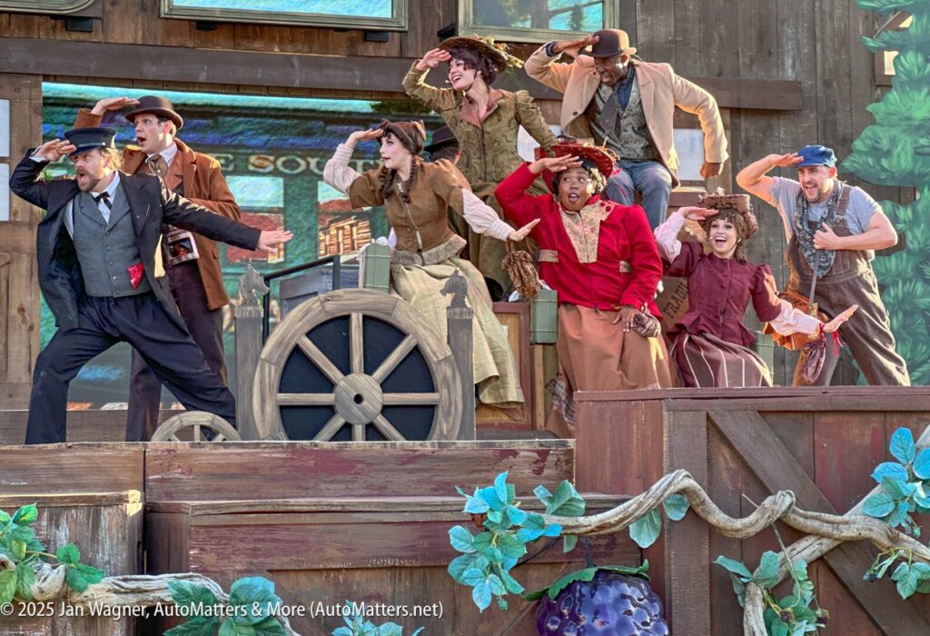 A group of performers in period costumes pose on an outdoor stage, with some standing on a wooden wagon and all shading their eyes as if looking into the distance.