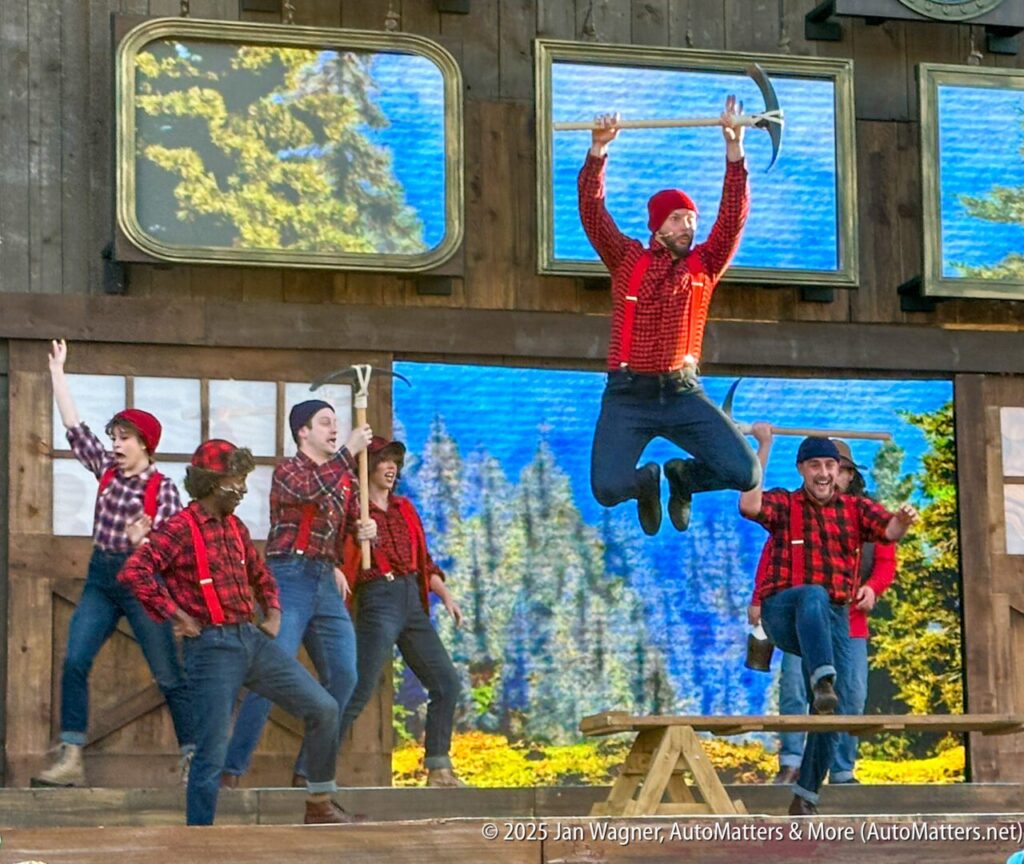 A group of performers in plaid shirts and suspenders, with one person mid-air jumping over a saw on stage, in front of a forest backdrop.