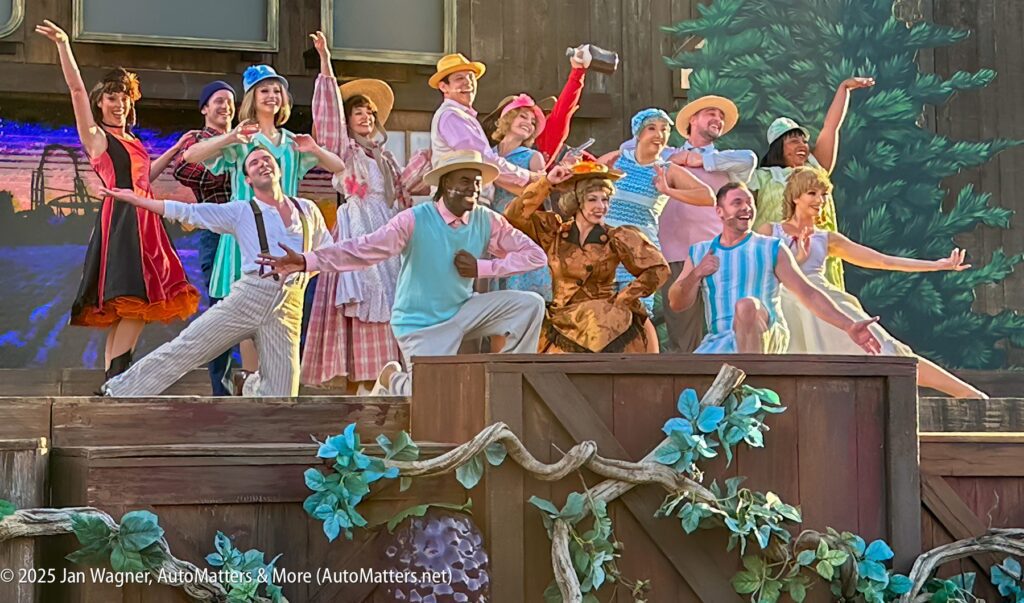 A group of performers in colorful costumes pose and smile on an outdoor stage decorated with vines and wooden crates.