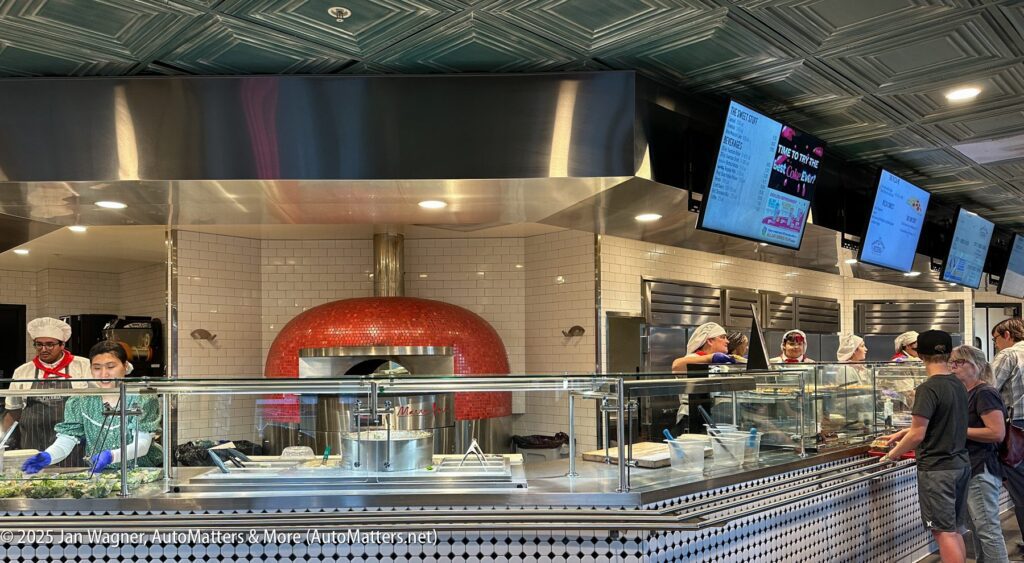 Several workers prepare food behind a glass counter in a modern pizzeria, with a red pizza oven and digital menu screens visible overhead. Customers wait in line at the counter.