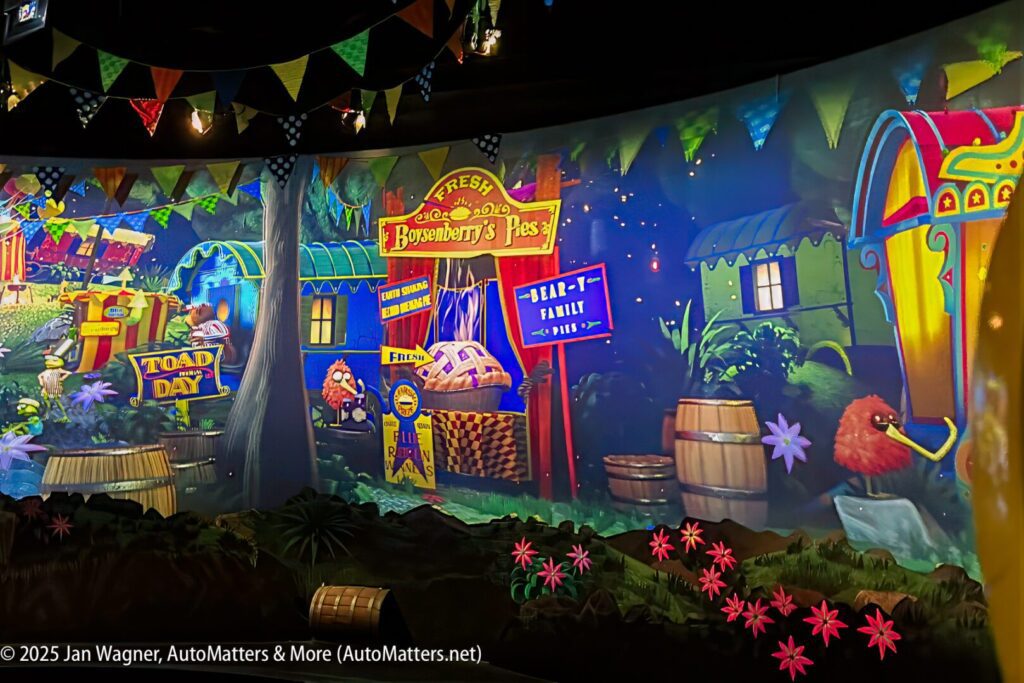 Brightly colored, animated scene of a whimsical village with market stalls, barrels, flowers, and festive flags, likely from an amusement park or themed attraction.
