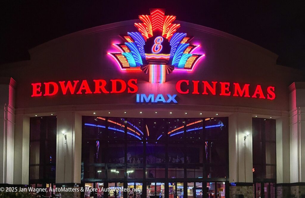 A brightly lit Edwards Cinemas IMAX theater entrance at night, featuring neon signage and reflections on the glass doors.
