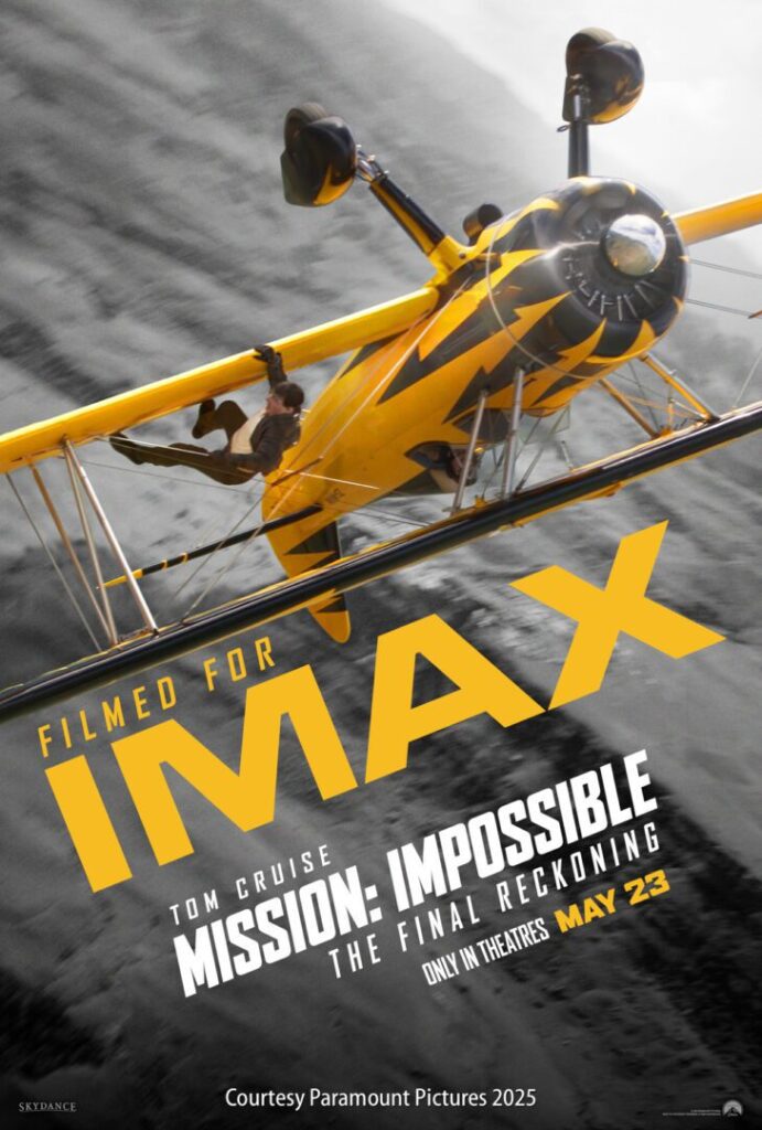A man hangs from the wing of a yellow biplane in flight, promoting "Mission: Impossible – The Final Reckoning," filmed for IMAX and releasing in theaters May 23.