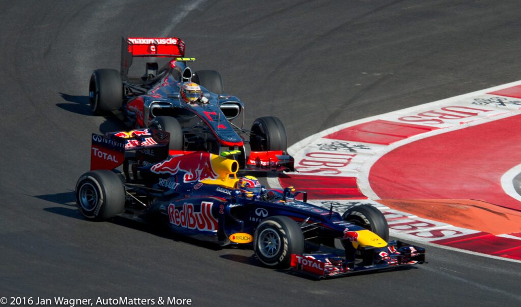 Two Formula 1 cars navigate a sharp corner on a racetrack, with one car closely following behind the other. Racing advertisements and curbs are visible on the track.