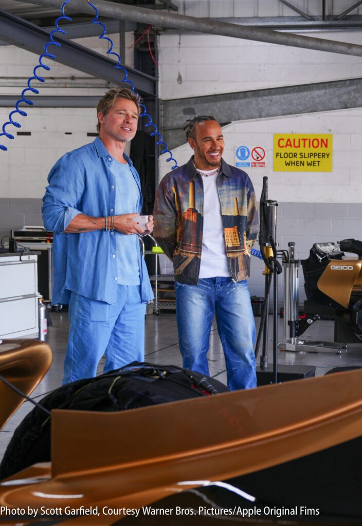 Two men stand and talk in a garage with technical equipment and a caution sign visible on the wall behind them.