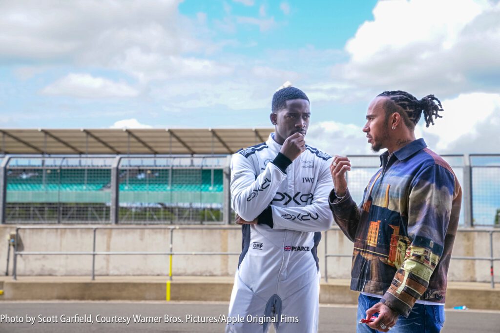Two men stand by a racetrack; one wears a white racing suit, the other wears a patterned jacket. They appear to be having a focused conversation.