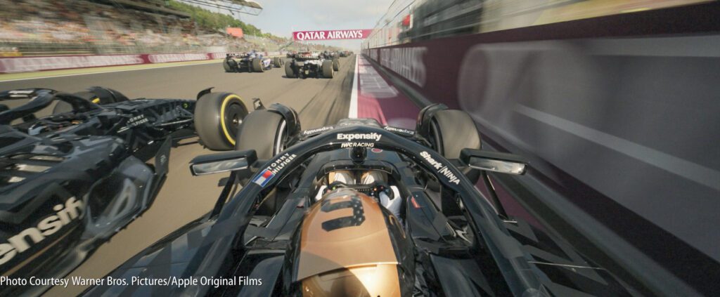 A Formula 1 car races at high speed on a track, closely followed by several other cars, with blurred motion and a crowd in the background.