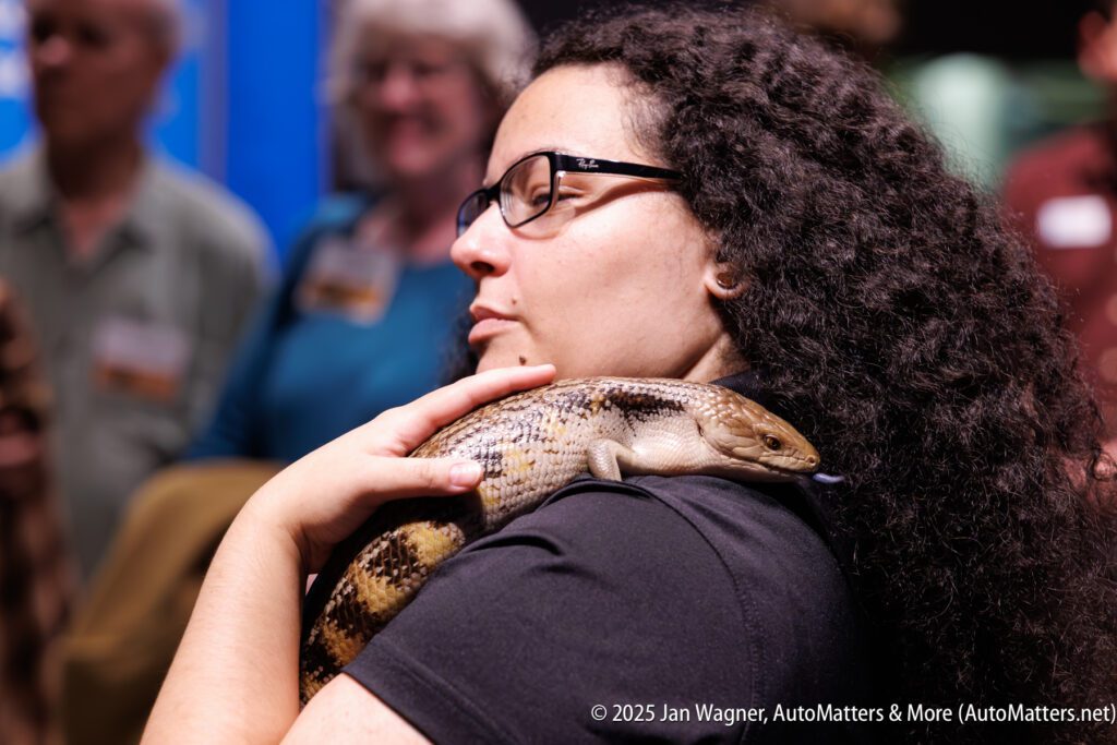 A woman with curly hair and glasses holds a large lizard on her shoulder while standing in a group of people indoors.