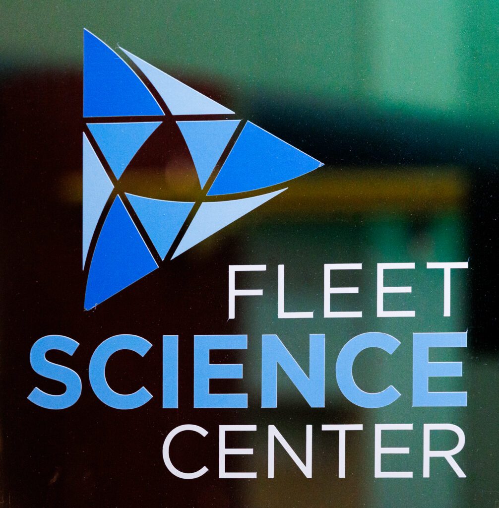 Fleet Science Center logo with a geometric blue triangle design and the words "FLEET SCIENCE CENTER" in white capital letters on a glass surface.